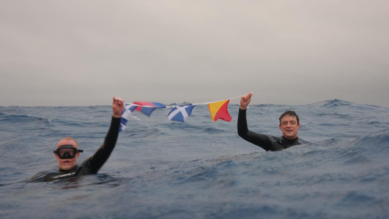 Two swimmers hold nautical signal flags in the open ocean near Point Nemo, Earth's oceanic pole of inaccessibility.