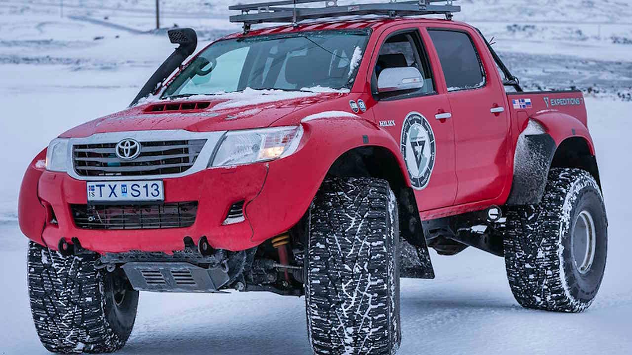 Red Toyota Hilux truck with 44 inch tires, snorkel and roof rack parked on a flat snowy surface.