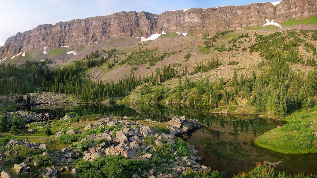 Scenic view of the Devil's Causeway ridgeline in the Flat Tops Wilderness, featuring a serene lake below the ridge, pine trees and a few remaining snow banks.