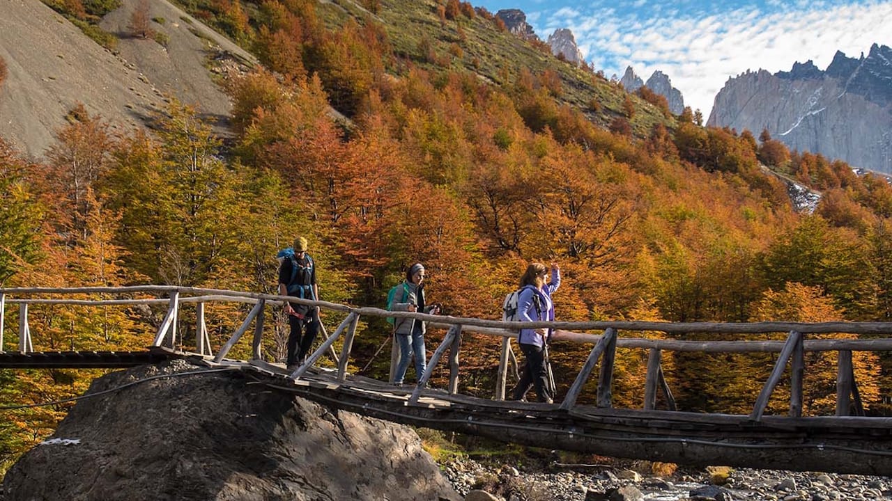 A group of hikers crosses a bridge over a river, framed by the majestic peaks of Torres del Paine National Park.
