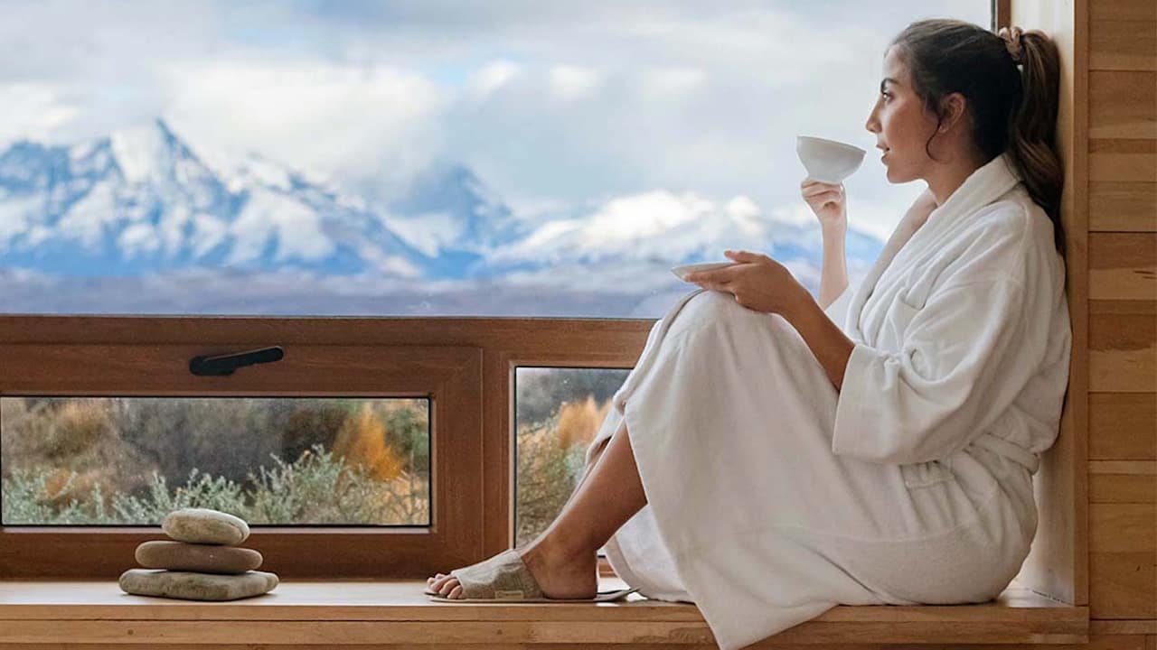 A woman relaxes in a robe on a window sill, enjoying coffee and the breathtaking view of Torres del Paine outside.