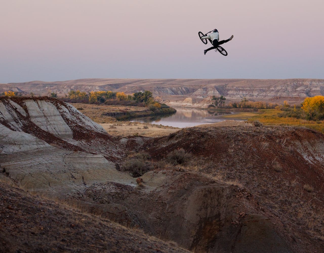A mountain biker jumping his bike with a river and yellow leafed trees in the background.