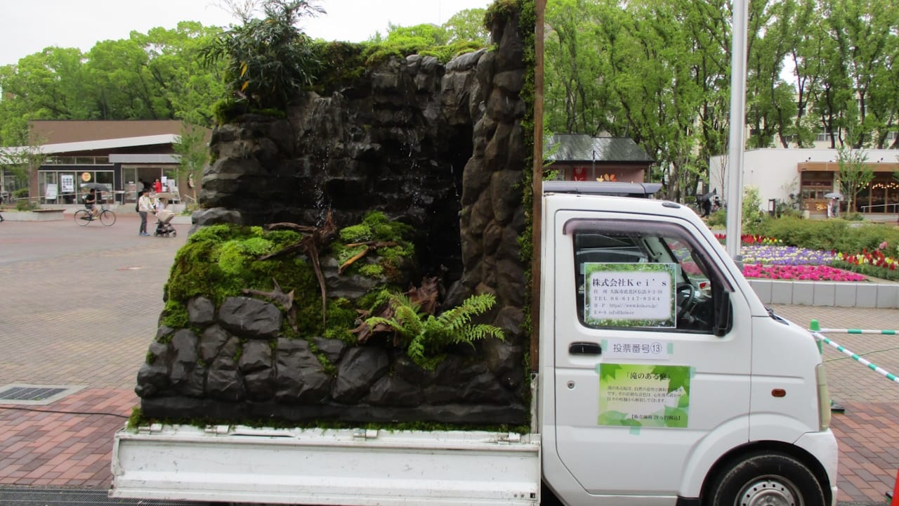 A tall rock face with water pouring down it into a garden area in the back of a small truck.