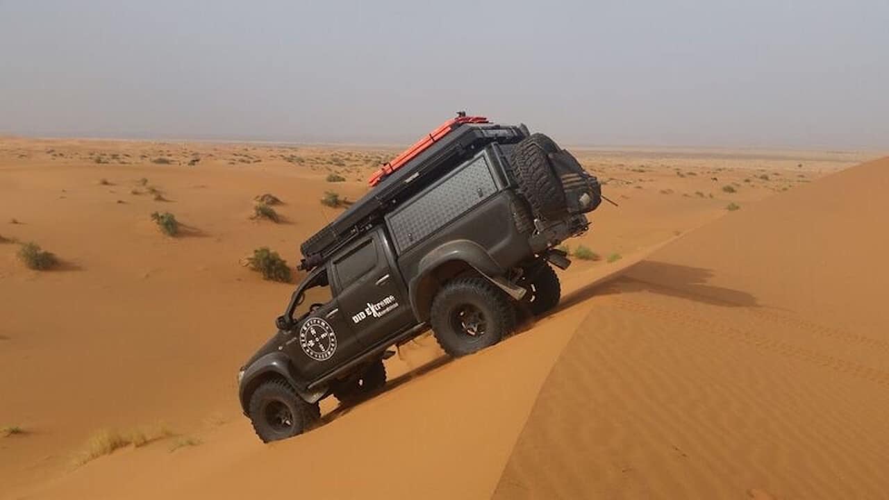 Black Toyota Hilux truck with cab and desert expedition gear shown driving down the steep side of a sand dune in the desert.