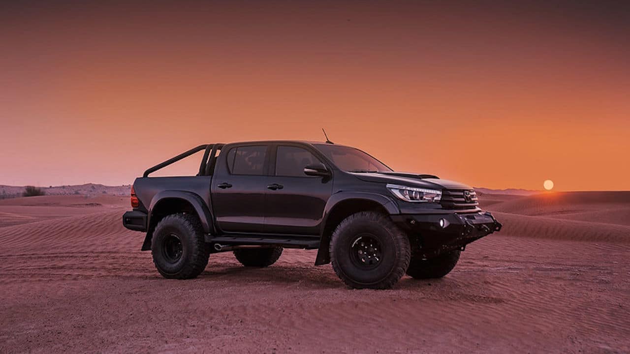 Toyota Hilux four door truck with large tires parked on desert sand with the sun setting behind.