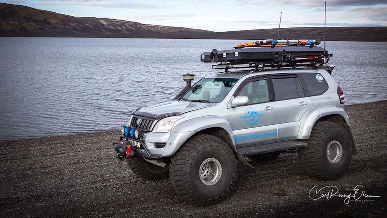 Silver Toyota Land Cruiser 4x4 SUV with huge tires and camping roof rack driving on the rocky shore of a lake.