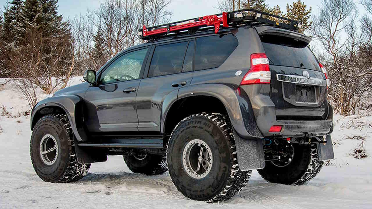 Black Land Cruiser on 44 inch tires with roof rack and red jack attached to it on a snowy surface.