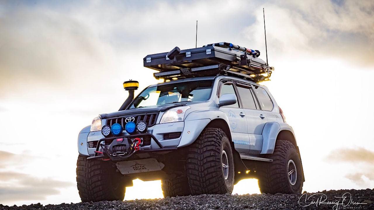White Toyota Land Cruiser with 46 inch tires parked on a rocky hill with a bright sunrise behind it.