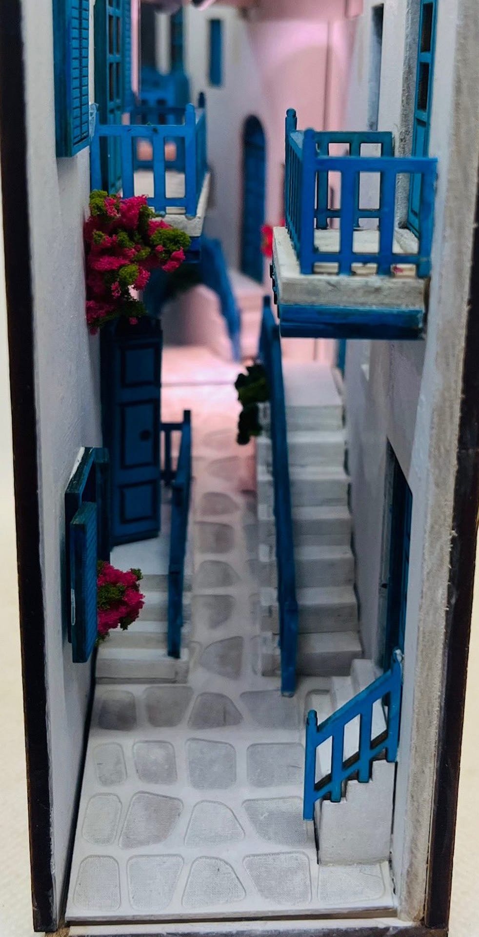 Book nook by Monde depicting a narrow whitewashed residential alley from a Greek island with blue trim and red flowers.